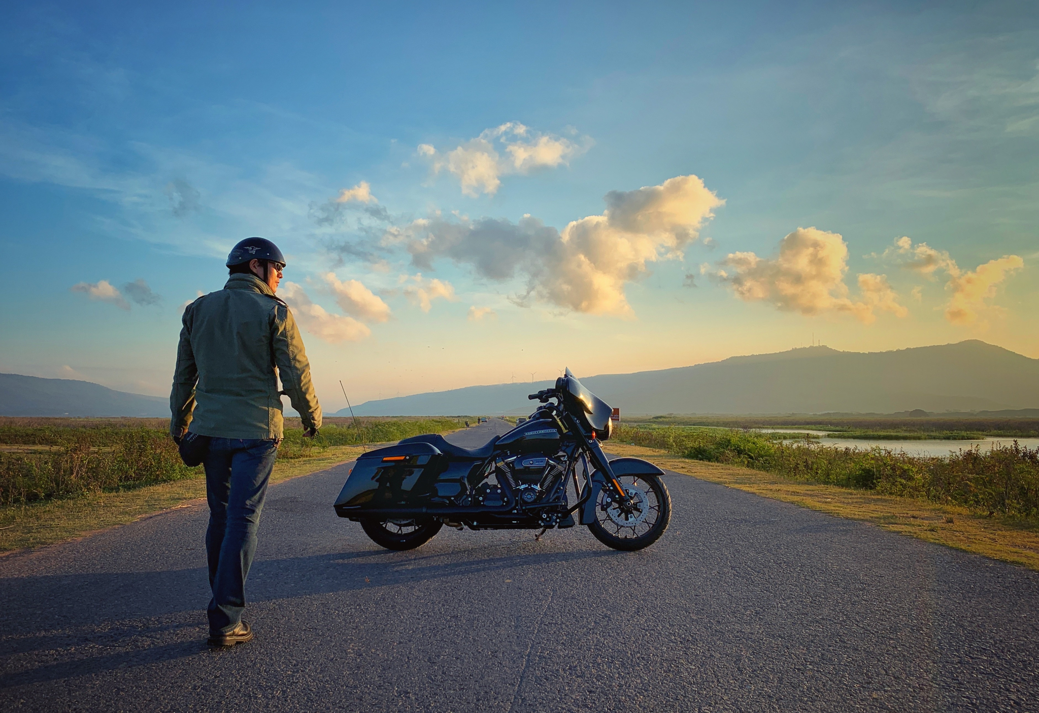 นักขี่สวมหมวกกะลาและแว่นตาขณะพักข้างรถ Harley-Davidson บนเส้นทางกลางแจ้ง